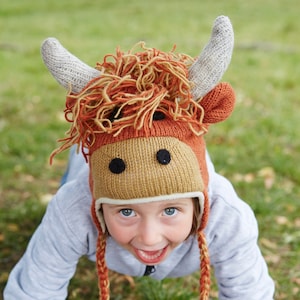 May include: A child wearing a brown and white knitted hat shaped like a cow with fuzzy brown hair. The hat has black eyes and a white nose. The child is wearing a light blue sweater and is looking at the camera with a big smile.