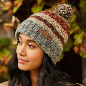 Könnte beinhalten: Eine gestrickte Mütze in Grau, Braun und Rot mit einem Bommel oben. Die Mütze hat einen braunen Leder-Patch mit dem Text "Pachamama" darauf.