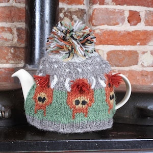 May include: A grey knitted teapot cosy with a pom-pom on top. The cosy features a green and orange pattern with a design of three stylised animals. The cosy is sitting on top of a black stove.