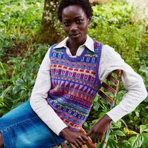 Op de afbeelding: Een vrouw draagt een wit shirt en een kleurrijk gebreid vest en zit op een houten stoel in een tuin. Het vest heeft een geometrisch patroon in tinten van paars, oranje, blauw en groen.