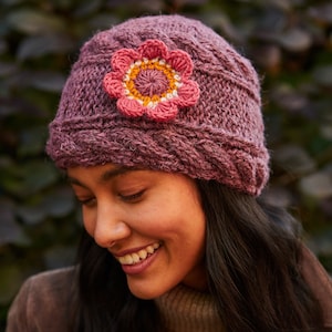 Puede incluir: Una mujer lleva un gorro de punto morado con una flor de ganchillo rosa y amarilla en un lado. Está sonriendo y mirando hacia abajo.