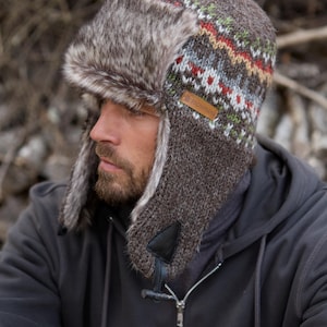May include: A brown trapper hat with faux fur ear flaps and a patterned knit crown. The hat features a leather patch with text. The hat is designed to provide warmth and protection from the cold.