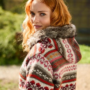 May include: A hooded knit sweater featuring a multi-colored fair isle pattern in shades of red, cream, brown, and pink. The garment includes a hood trimmed with dark grey and brown faux fur, long sleeves, and a thick, textured wool-like knit construction.