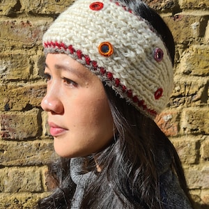 Op de afbeelding: Een crèmekleurige gebreide hoofdband met een dikke, grove garentextuur. De hoofdband is voorzien van een decoratieve rode gestikte rand langs de onderkant en diverse kleine, ronde, meerkleurige geborduurde versieringen.