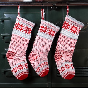 May include: Three red and white knitted Christmas stockings hanging on a black cast iron stove. The stockings have a snowflake pattern.