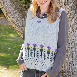 May include: A gray sleeveless sweater with a green and purple thistle pattern. The sweater has a round neckline and is worn over a gray long-sleeved shirt. The person is wearing a headband with a similar thistle pattern.