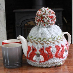 以下が含まれることがあります： 白いポンポンが付いたニットのティーポットカバー。カバーは、グレーの背景に赤いリスと緑の葉の模様で飾られています。