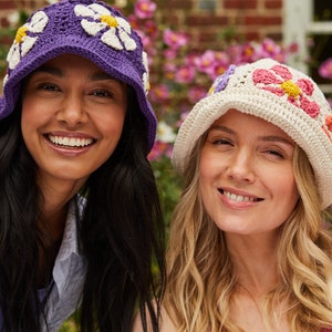 May include: Two women wearing crocheted flower hats. The woman on the left is wearing a purple hat with white flowers. The woman on the right is wearing a white hat with pink, orange, and yellow flowers.