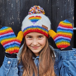 May include: A young person wearing a white knit hat with a rainbow heart design and a rainbow striped knit mitten on each hand. The hat has a white pom-pom on top.