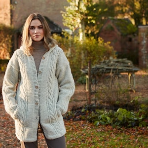 May include: A woman wearing a light gray hooded cardigan with a cable knit pattern and pockets. The cardigan has a button closure and is worn over a brown turtleneck sweater.