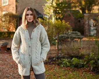 May include: A woman wearing a light gray hooded cardigan with a cable knit pattern and pockets. The cardigan has a button closure and is worn over a brown turtleneck sweater.