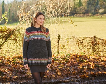 May include: A dark gray sweater with a colorful geometric pattern across the chest and at the hem. The sweater is worn with a dark skirt and tights. The background features a field, trees, and a fence, suggesting an outdoor setting.