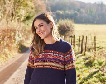 May include: A woman wearing a blue sweater with a colorful patterned design stands on a dirt road. The sweater has a fair isle pattern with red, orange, yellow, green, blue, and purple colors. The woman is smiling and looking to the right.