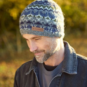 May include: A knitted beanie in shades of blue, green, and white, with a small brown leather tag that reads "PACHAMAMA". The hat is worn by a person wearing a denim jacket.