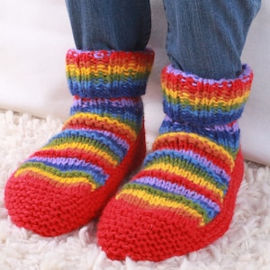 Op de afbeelding: Een paar rode gebreide pantoffels met regenboogstrepen. De sokken worden gedragen door iemand die een blauwe jeans draagt.