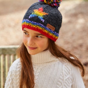 May include: A gray knitted beanie with a rainbow star design and a colorful pom pom on top. The beanie has a red, orange, yellow, green, blue, and purple stripe around the bottom edge. The beanie has a brown leather label with the text "Pachamama" on it.