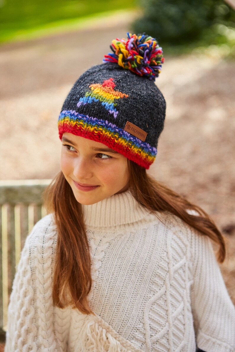 May include: A gray knitted beanie with a rainbow star design and a colorful pom pom on top. The beanie has a red, orange, yellow, green, blue, and purple stripe around the bottom edge. The beanie has a brown leather label with the text "Pachamama" on it.