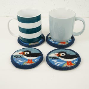 Puede incluir: Cuatro posavasos redondos de fieltro con fondo azul y un diseño de frailecillo blanco y naranja. Dos tazas están encima de los posavasos. Una taza es blanca con un diseño de rayas azules y la otra taza es azul claro.