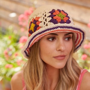 Op de afbeelding: Een gehaakte bucket hat met een beige basis en veelkleurige granny square motieven in paars, oranje en groen. De rand is voorzien van horizontale strepen in paars, oranje en beige, wat zorgt voor een getextureerd en handgemaakt uiterlijk.