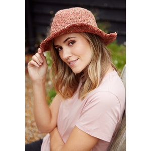 May include: A woman wearing a pink t-shirt and a crocheted sun hat with a wide brim. The hat is a mix of red and brown colors.