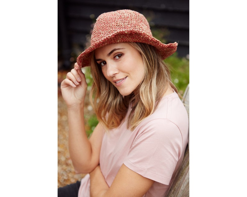 May include: A woman wearing a pink t-shirt and a crocheted sun hat with a wide brim. The hat is a mix of red and brown colors.