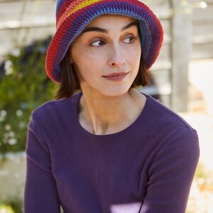 May include: A woman wearing a purple long-sleeved shirt and a colorful crocheted bucket hat with rainbow stripes.