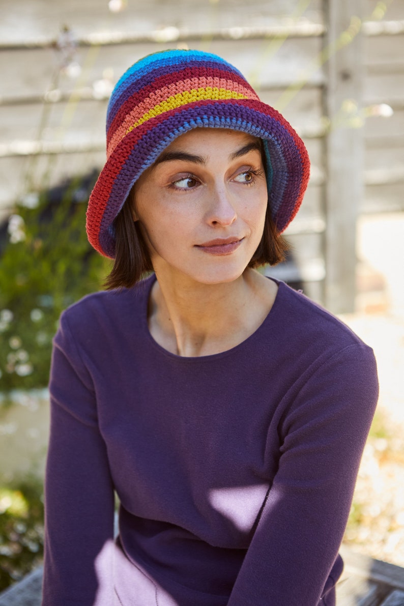May include: A woman wearing a purple long-sleeved shirt and a colorful crocheted bucket hat with rainbow stripes.