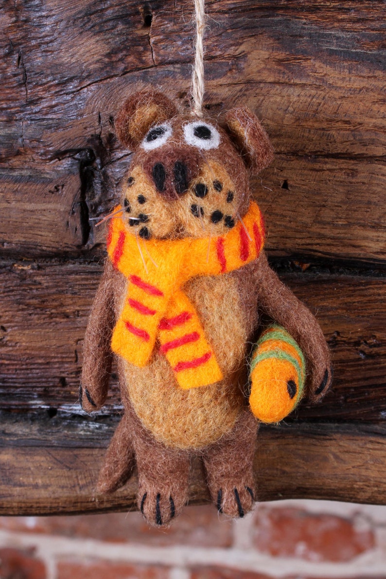 May include: A brown felt animal with a yellow and orange striped scarf and a yellow felt ball in its paw. The animal is hanging from a string against a wooden background.
