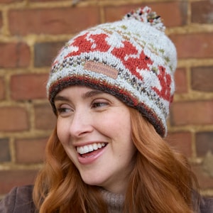 May include: A woman wearing a grey and red knitted hat with a pom pom on top. The hat has a brown leather label with the text "Pachamama" on it.