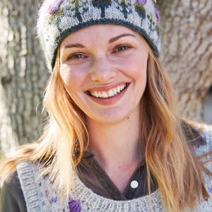 May include: A woman wearing a gray and green knitted headband with purple flowers and a gray knitted vest with purple flower embroidery.