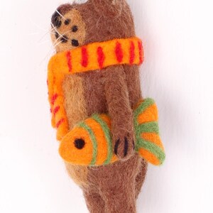 May include: A brown felt animal ornament with a red and orange striped scarf and a green and orange fish in its paw.
