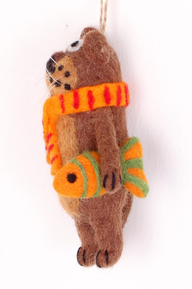 May include: A brown felt animal ornament with a red and orange striped scarf and a green and orange fish in its paw.