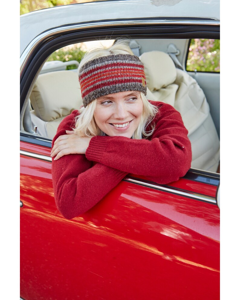 May include: A woman wearing a red sweater and a striped knit headband looks out the window of a red car.