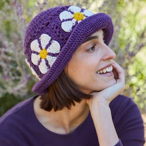 May include: A purple crochet hat with white and yellow daisy flowers. The hat has a wide brim and is made with a crocheted stitch.