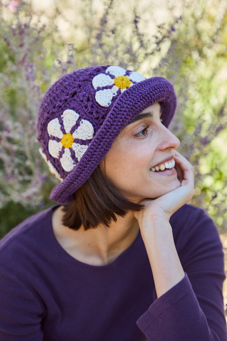 May include: A purple crochet hat with white and yellow daisy flowers. The hat has a wide brim and is made with a crocheted stitch.