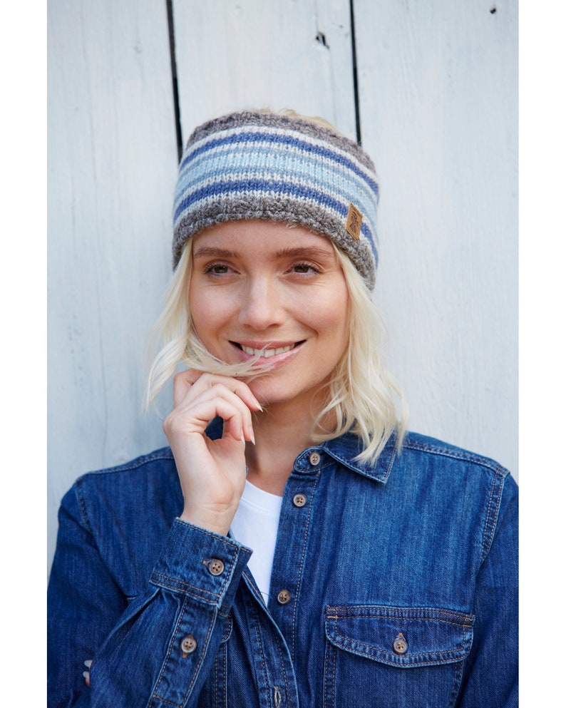 May include: A blue and gray striped knit headband with a brown leather tag. The headband is worn by a person with blonde hair and a denim shirt.