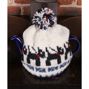 May include: White knitted teapot cozy with a black and white dog pattern and a red pom pom on top. The cozy is on a blue teapot.