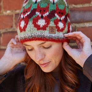 May include: A red, green, and white knit hat with a pom-pom on top. The hat features a repeating pattern of stylized foxes. The hat has a brown leather tag with the text "El Sol" on it.