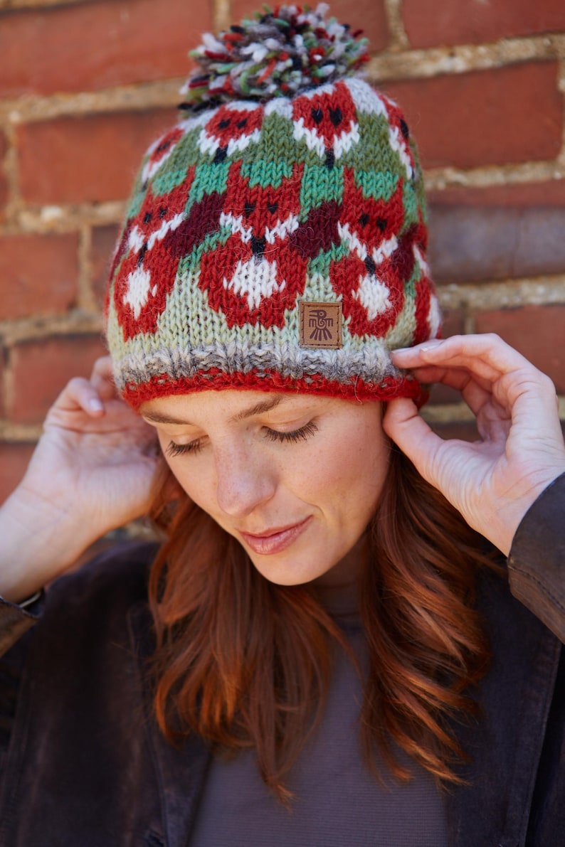May include: A red, green, and white knit hat with a pom-pom on top. The hat features a repeating pattern of stylized foxes. The hat has a brown leather tag with the text "El Sol" on it.