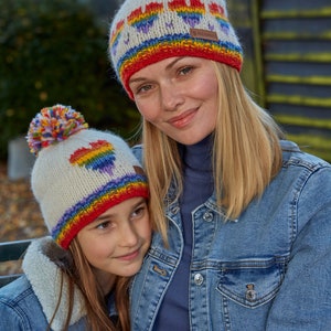 May include: Two people wearing white knit hats with rainbow hearts and pom poms. The hats have a red, orange, yellow, green, blue, indigo, and violet stripe. The people are wearing denim jackets.