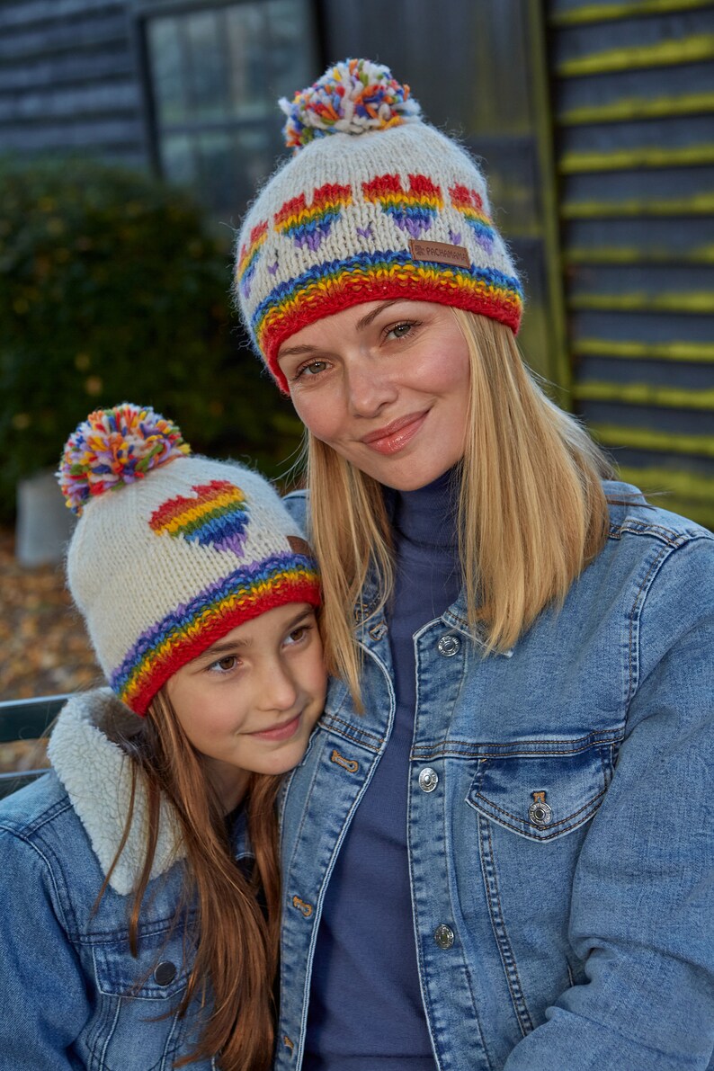 May include: Two people wearing white knit hats with rainbow hearts and pom poms. The hats have a red, orange, yellow, green, blue, indigo, and violet stripe. The people are wearing denim jackets.