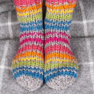 May include: A pair of colorful, hand-knitted socks. The socks feature horizontal stripes in shades of pink, orange, yellow, green, blue, and gray. The cuffs are a vibrant pink, and the toes are a solid gray. The socks are designed for warmth and comfort.