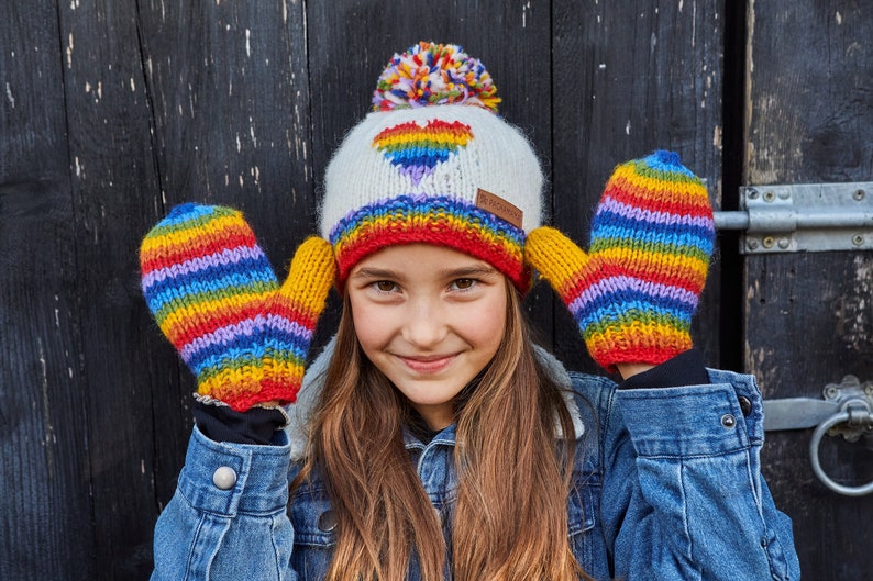May include: A young person wearing a white knit hat with a rainbow heart design and a rainbow striped knit mitten on each hand. The hat has a white pom-pom on top.