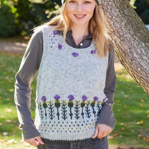 May include: A gray sleeveless sweater with a green and purple thistle pattern. The sweater has a round neckline and is worn over a gray long-sleeved shirt. The person is wearing a purple and green headband with a thistle pattern.