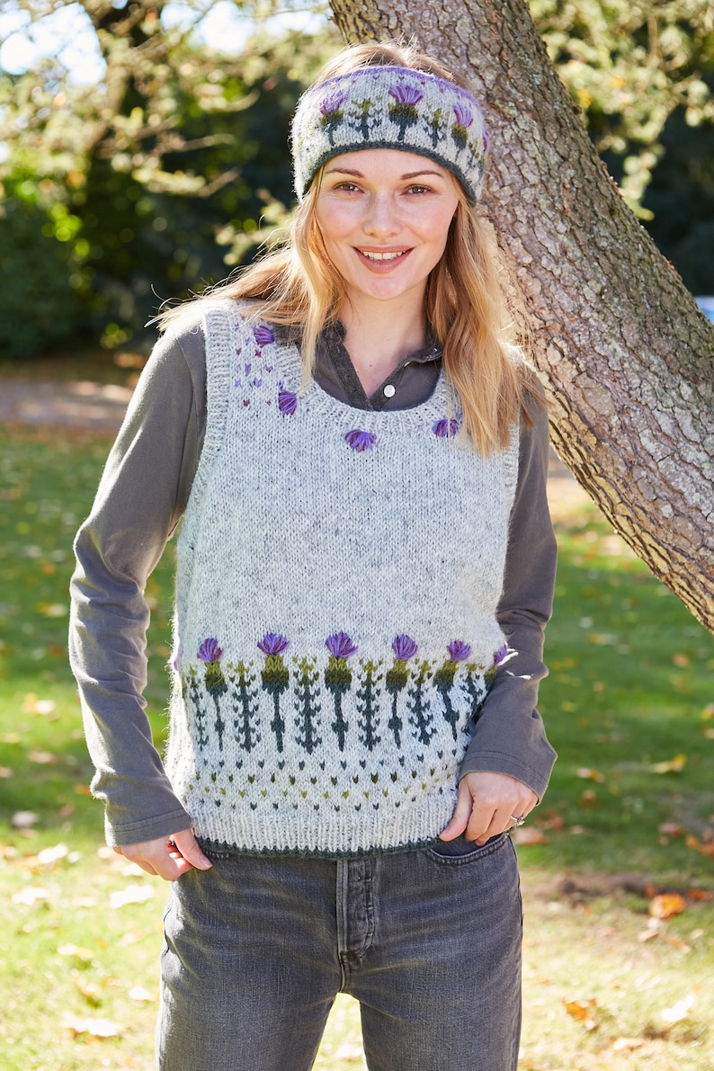 May include: A gray sleeveless sweater with a green and purple thistle pattern. The sweater has a round neckline and is worn over a gray long-sleeved shirt. The person is wearing a purple and green headband with a thistle pattern.