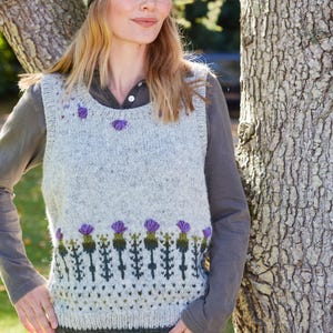 May include: A gray knitted vest with a purple and green thistle pattern. The vest has a scoop neck and is sleeveless. The model is wearing the vest over a gray shirt and blue jeans. The model is also wearing a purple and green knitted headband with a thistle pattern.