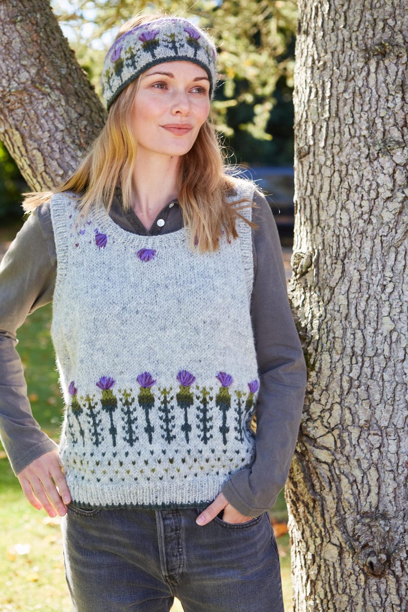 May include: A gray knitted vest with a purple and green thistle pattern. The vest has a scoop neck and is sleeveless. The model is wearing the vest over a gray shirt and blue jeans. The model is also wearing a purple and green knitted headband with a thistle pattern.