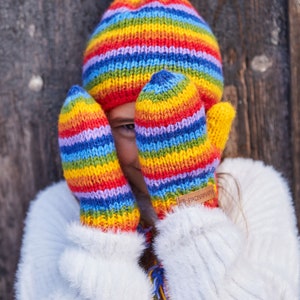 May include: A child wearing a white fuzzy sweater and rainbow striped knit hat and mittens. The mittens have a brown leather tag with the text "Pachamama" on them.