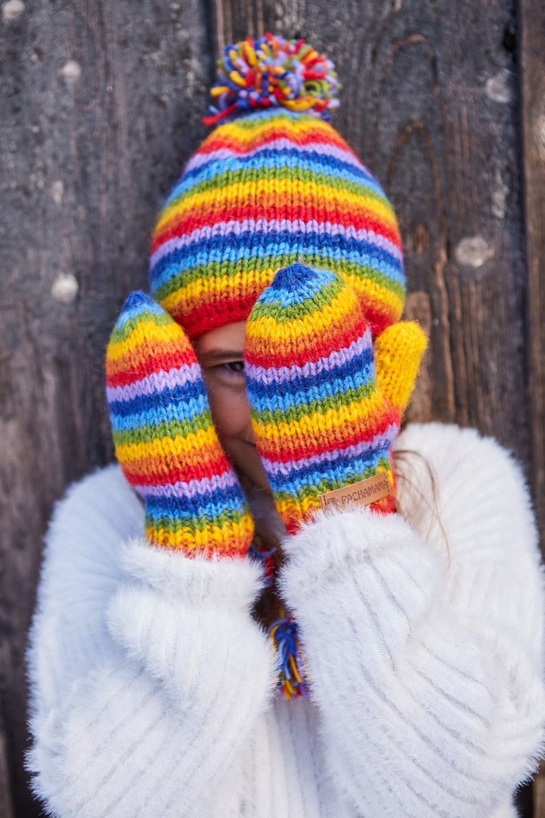 May include: A child wearing a white fuzzy sweater and rainbow striped knit hat and mittens. The mittens have a brown leather tag with the text "Pachamama" on them.