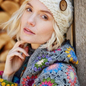 May include: A woman wearing a white crocheted headband with a wooden button and a grey crocheted sweater with colourful floral designs.